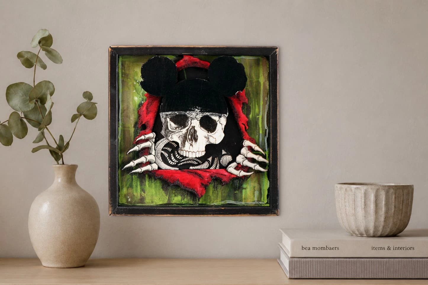 Schädel mit Mickey-Mouse-Ohren und roten Tüchern vor grünem Hintergrund, als Kunstwerk an der Wand eines hellen Raums.
