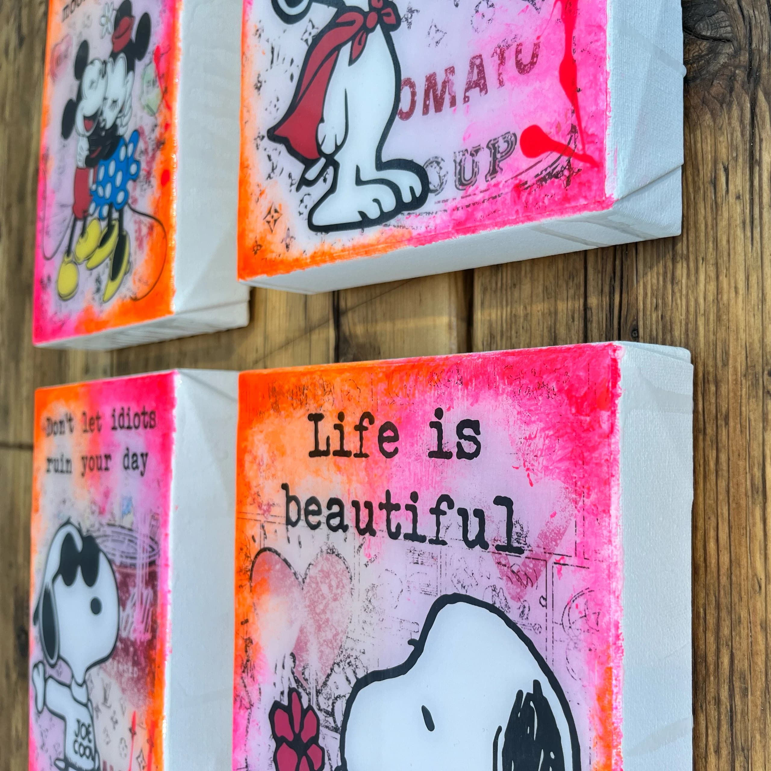 Mehrere Snoopy-Bilder auf Leinwand hängen vor einer Holzvertäfelung, eines mit dem Text „Life is beautiful“.