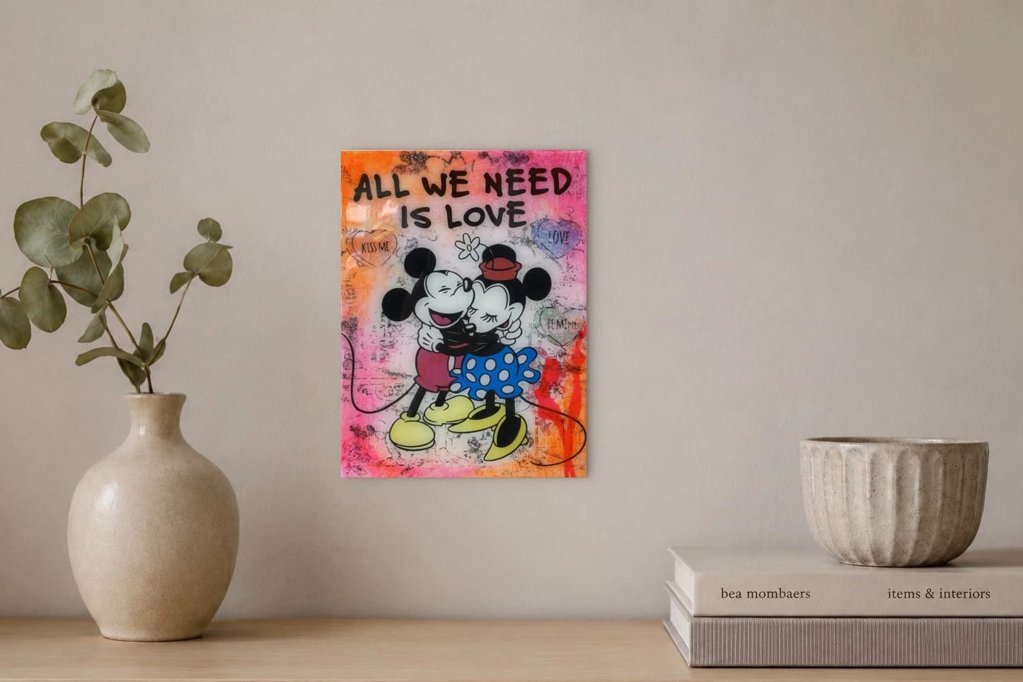 Mickey und Minnie umarmen sich vor ALL WE NEED IS LOVE auf pinkem Hintergrund mit Herzen, als Kunstwerk an der Wand eines hellen Raums.