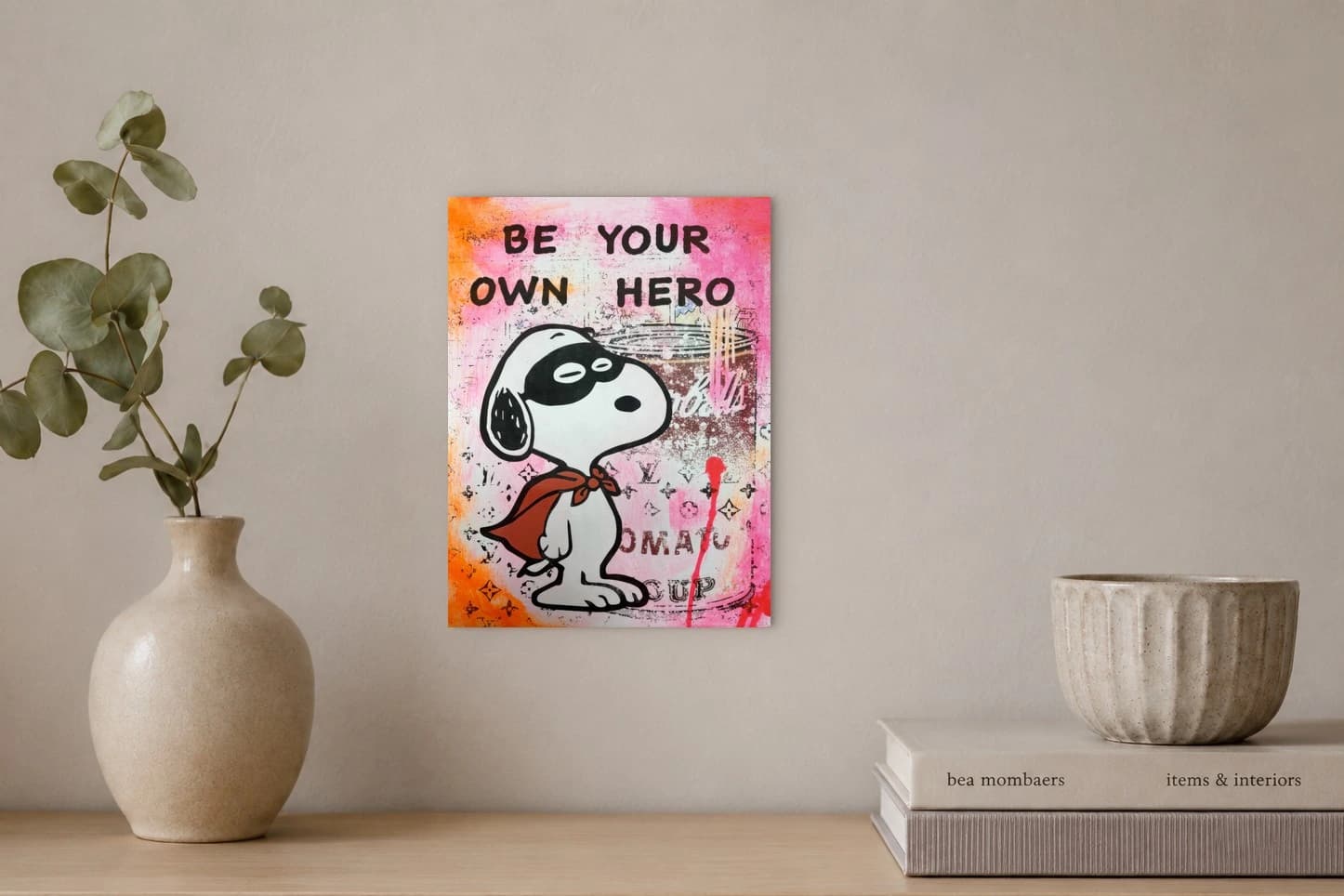 Snoopy mit rotem Halstuch vor pinkem Hintergrund und BE YOUR OWN HERO, als Kunstwerk an der Wand eines hellen Raums.