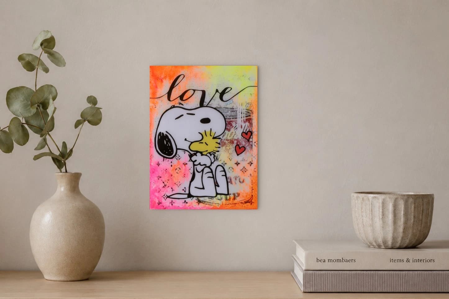 Snoopy küsst einen gelben Vogel vor buntem Hintergrund mit love und zwei roten Herzen, als Kunstwerk an der Wand eines hellen Raums.