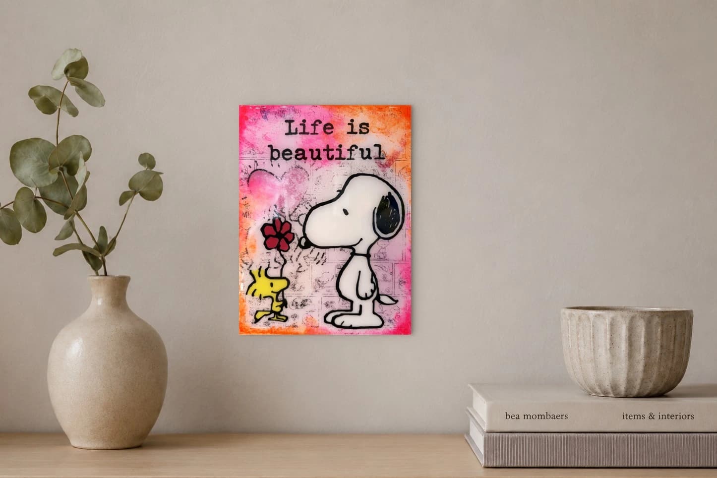 Snoopy steht neben Woodstock mit roter Blume und dem Schriftzug Life is beautiful auf rosa-orangefarbenem Grund, als Kunstwerk an der Wand eines hellen Raums.