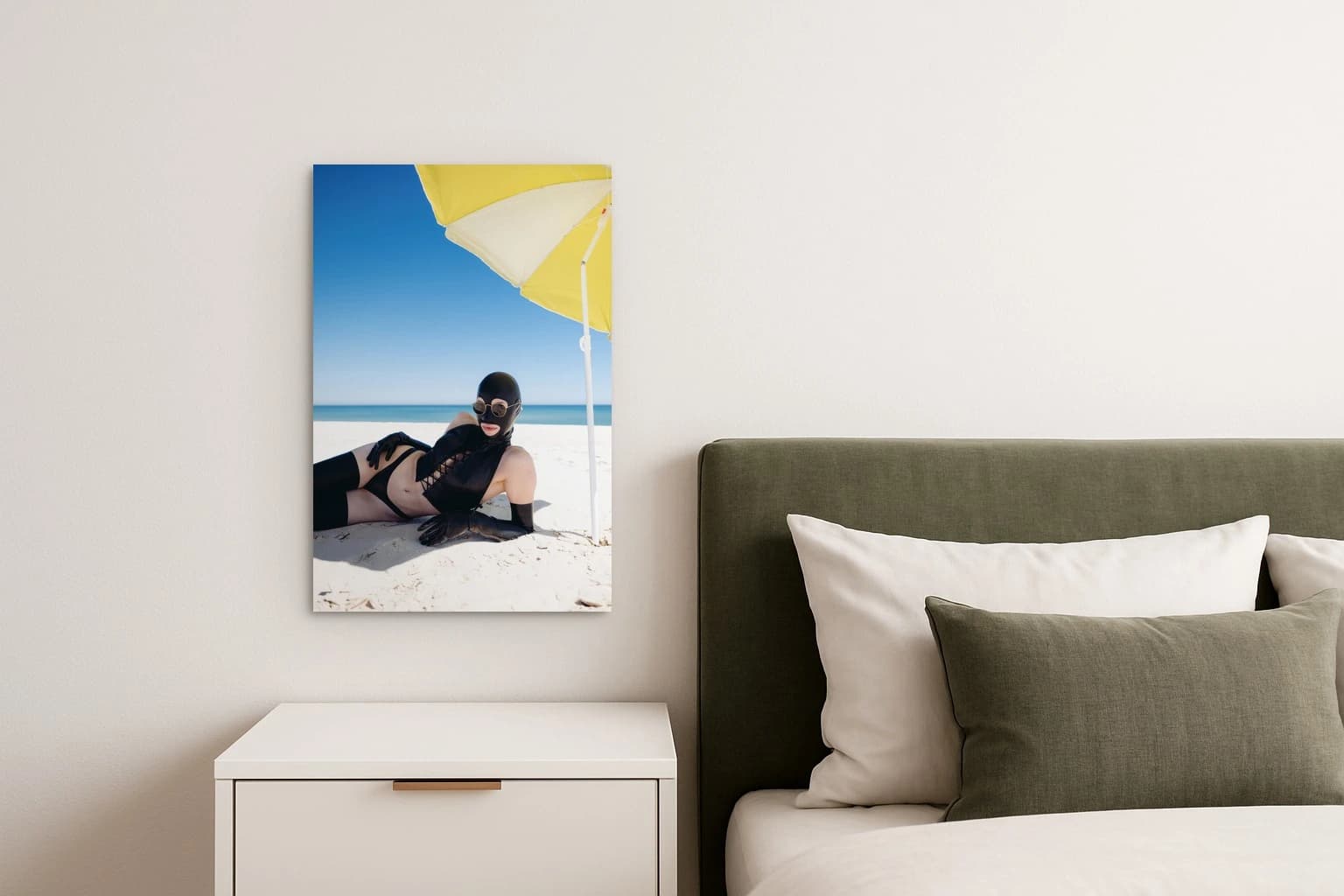 Eine Person in schwarzer Maske und Bademode liegt am Sandstrand unter einem gelben Sonnenschirm, als Kunstwerk an der Wand eines Schlafzimmers.