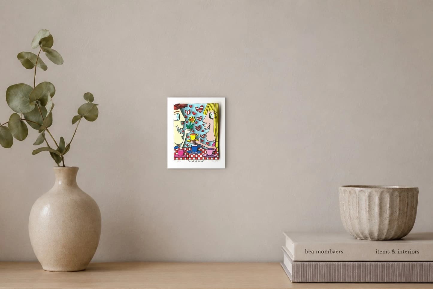 Zwei Gesichter sitzen am Tisch mit Kaffeetassen, Herzen und einer Blume vor blauem Hintergrund, als Kunstwerk an der Wand eines hellen Raums.