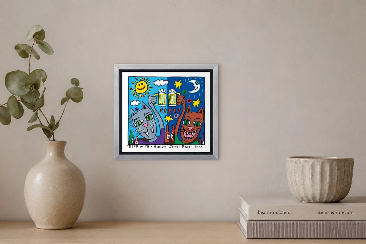 Gerahmtes Bild mit zwei Katzen, die Bierkrüge anstoßen, vor Sternen, Sonne, Mond und Flaschen, als Kunstwerk an der Wand eines hellen Raums.