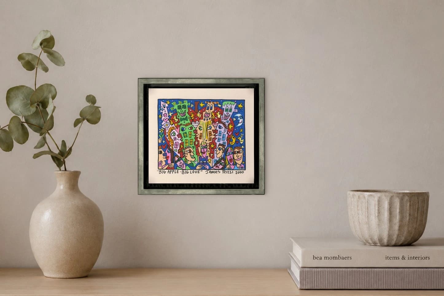 Gerahmtes Bild mit bunten Gesichtern, Sternen und Häusern auf Blau, darunter „Big Apple Big Love“, als Kunstwerk an der Wand eines hellen Raums.