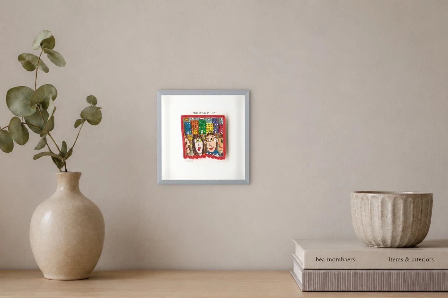 Gerahmtes Blatt mit roter Zeichnung von vier Gesichtern vor bunten Häusern und dem Text „DO DROP IN“, als Kunstwerk an der Wand eines hellen Raums.