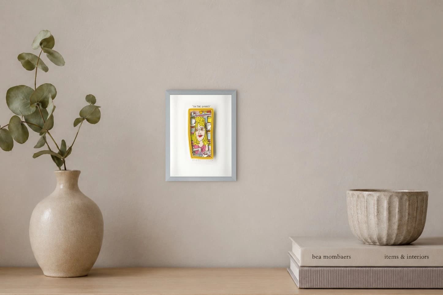 Gerahmtes Blatt mit gelbem Bild einer Frau zwischen Geschirr und dem Text „DO THE DISHES“, als Kunstwerk an der Wand eines hellen Raums.