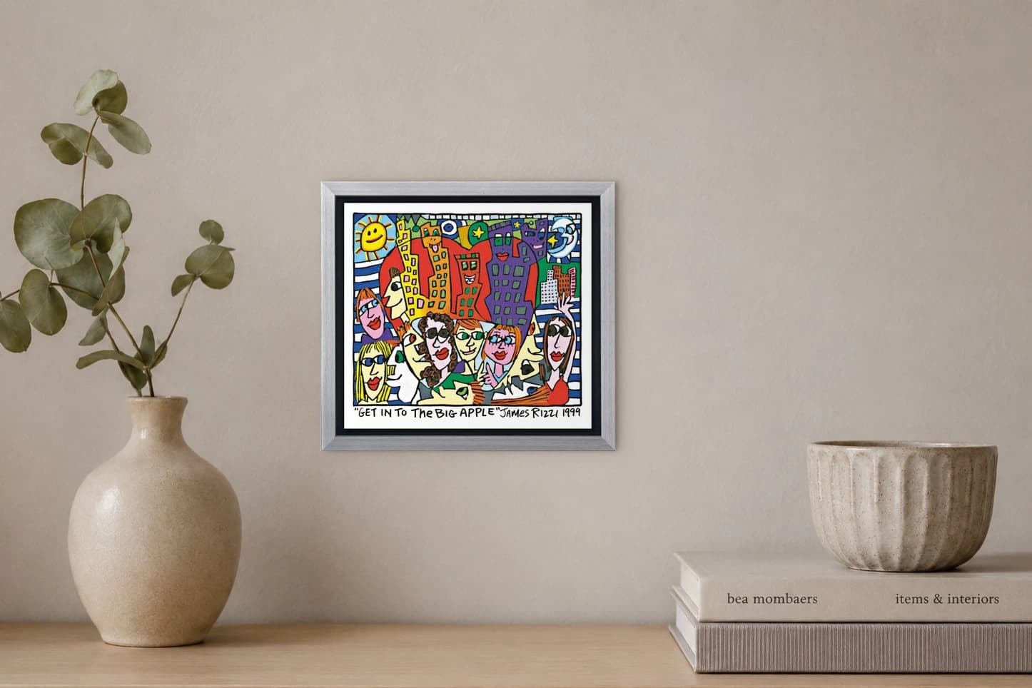 Gerahmtes Bild mit bunten Gesichtern, Hochhäusern, Sonne, Mond und Schriftzug „GET IN TO THE BIG APPLE“, als Kunstwerk an der Wand eines hellen Raums.