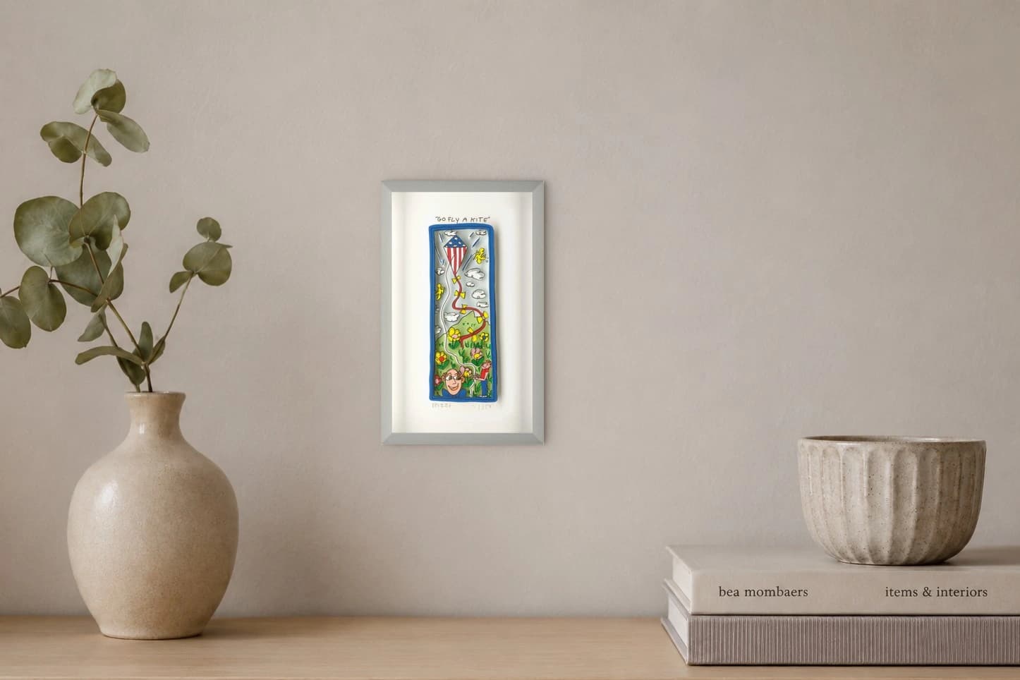 Gerahmtes Bild mit Go Fly a Kite, rotem weißem Drachen, Wolken und Figuren auf einer Wiese, als Kunstwerk an der Wand eines hellen Raums.