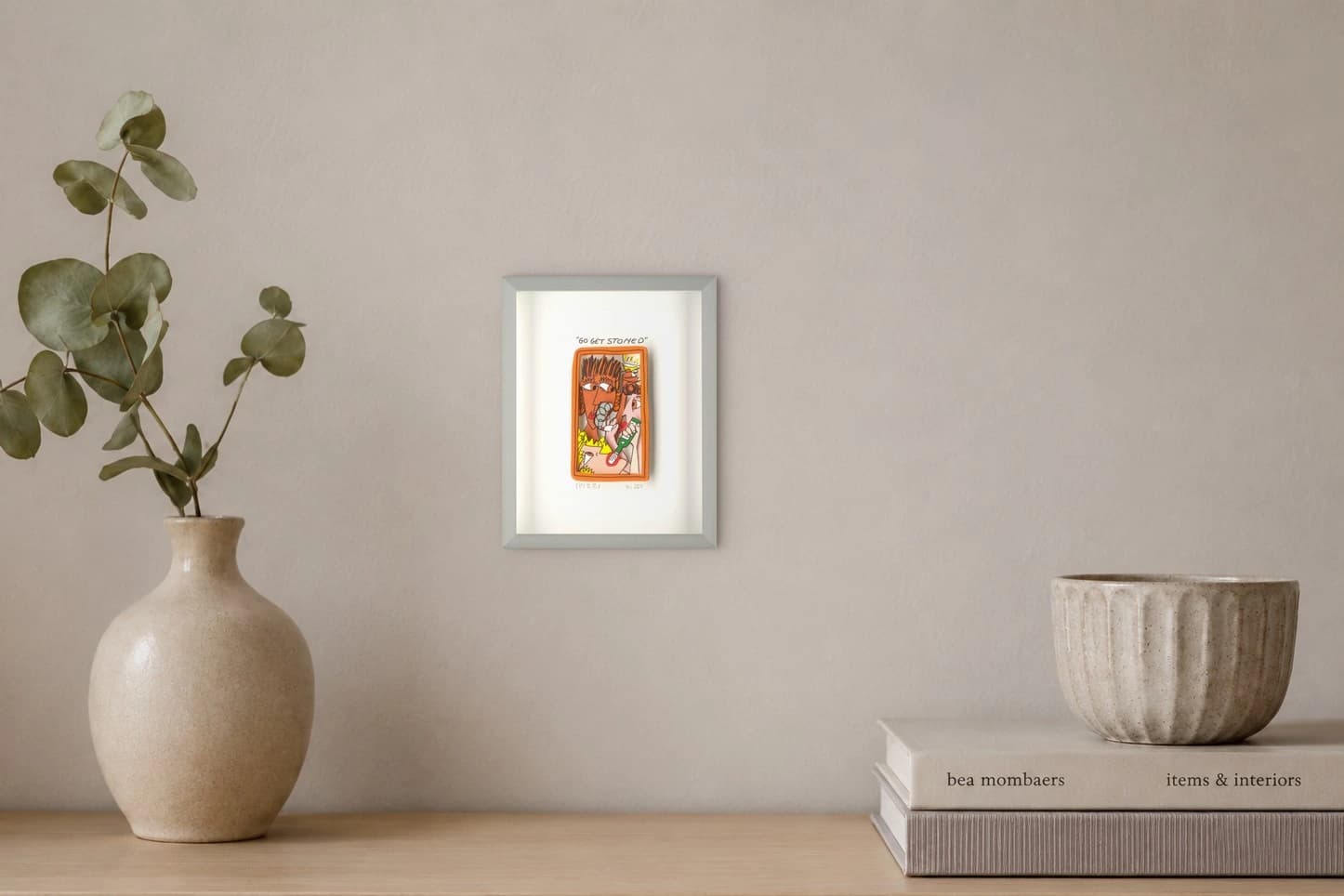 Gerahmtes Blatt mit orangefarbenem Bild und Schriftzug „GO GET STONED“, als Kunstwerk an der Wand eines hellen Raums.
