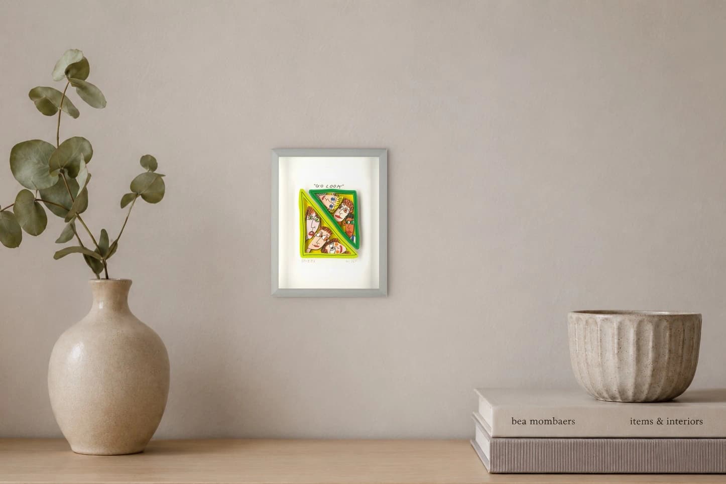 Gerahmtes Blatt mit buntem Popup-Motiv aus sechs Gesichtern und dem Schriftzug „GO LOOK“, als Kunstwerk an der Wand eines hellen Raums.