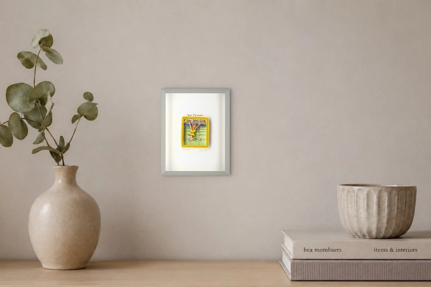 Gerahmtes Blatt mit kleiner Cheerleaderin in Gelb und „Go Team“, als Kunstwerk an der Wand eines hellen Raums.
