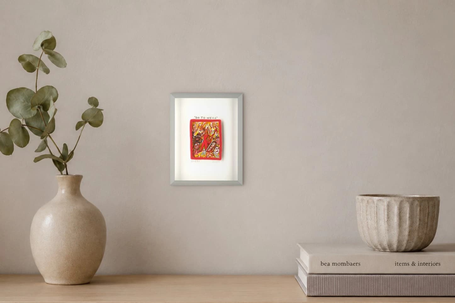 Gerahmtes Blatt mit rotem Bild und dem Text „GO TO HELL“ mit Figuren im Feuer, als Kunstwerk an der Wand eines hellen Raums.
