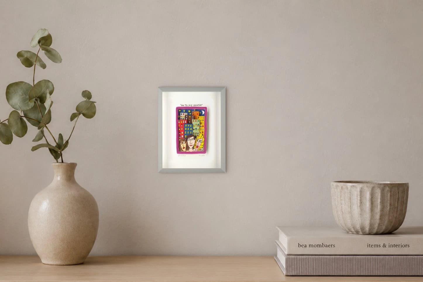 Farbige Häuser und drei Gesichter in einem pinken Rahmen mit „Go To My House“, als Kunstwerk an der Wand eines hellen Raums.