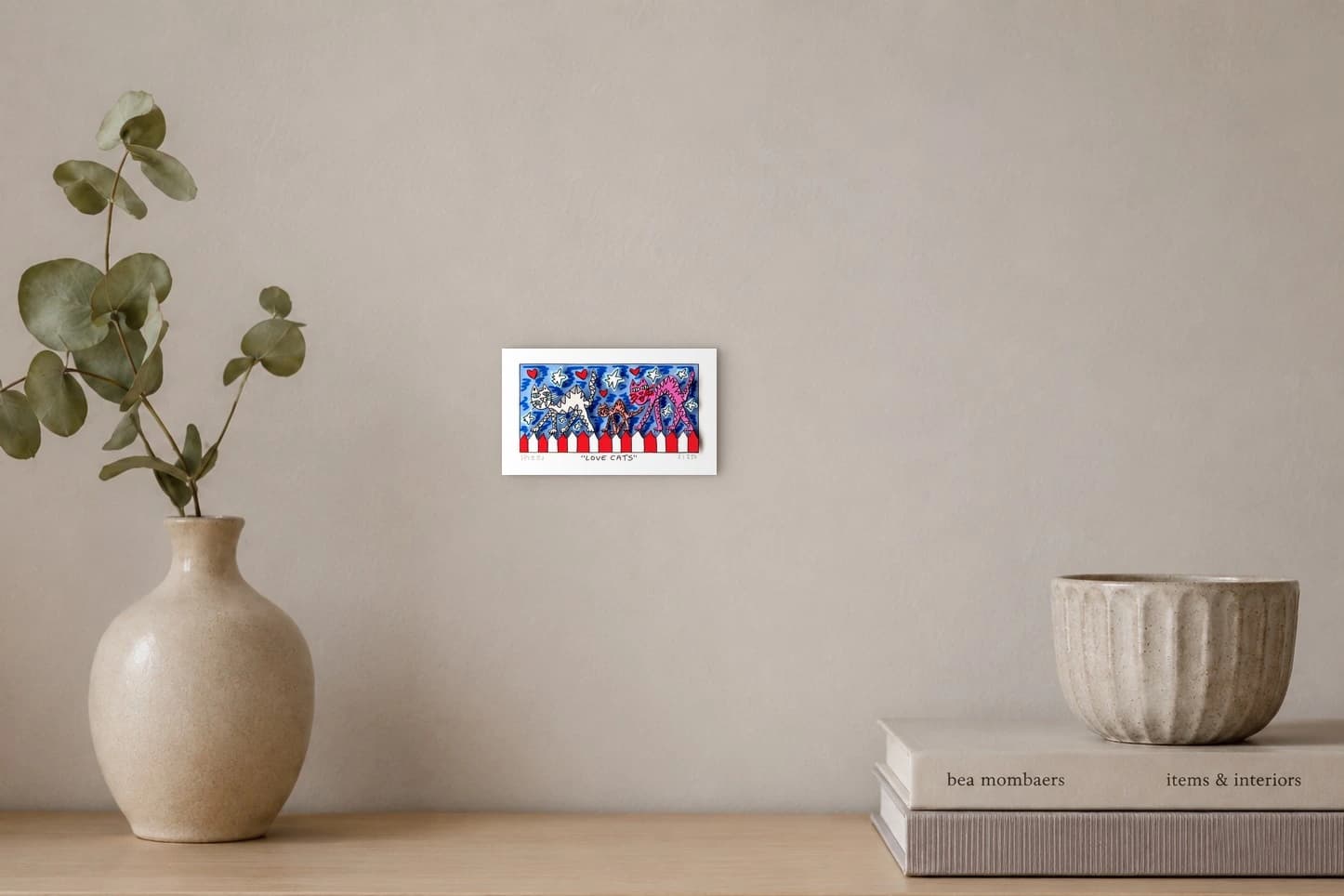 Drei Katzen vor blauem Hintergrund mit Herzen, Sternen und rotem-weißem Zickzackrand, als Kunstwerk an der Wand eines hellen Raums.