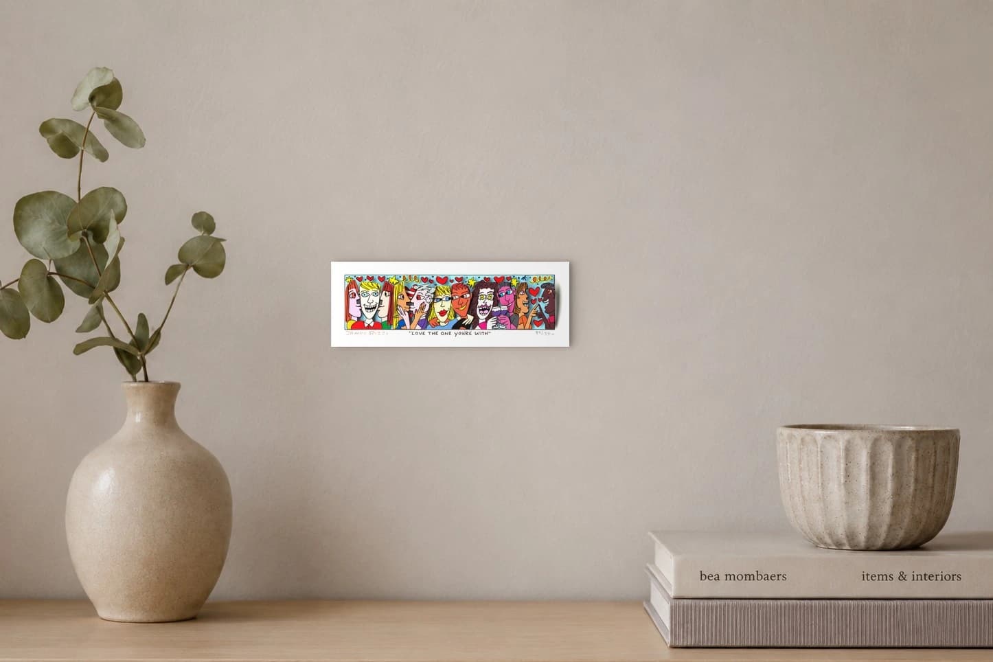Bunte Comicfiguren mit Herzen und Sternen, unten der Schriftzug "LOVE THE ONE YOU'RE WITH" und Signatur, als Kunstwerk an der Wand eines hellen Raums.