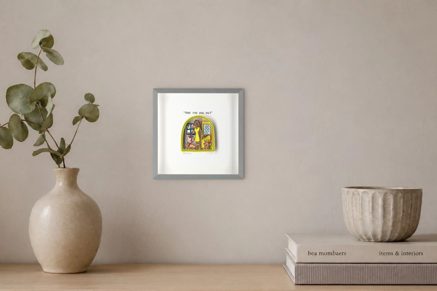 Gerahmtes Blatt mit gelber Zeichnung einer Frau und zwei Hunden sowie dem Text "TAKE THE DOG OUT", als Kunstwerk an der Wand eines hellen Raums.