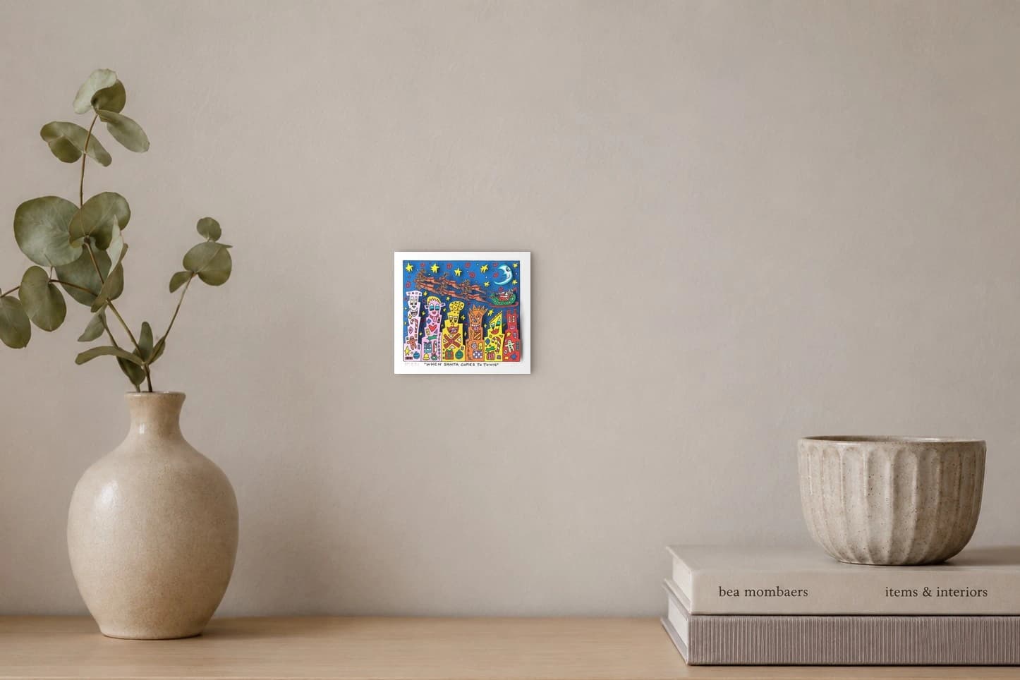 Santa im Schlitten mit Rentieren über bunten Häusern mit Gesichtern vor blauem Sternenhimmel, als Kunstwerk an der Wand eines hellen Raums.