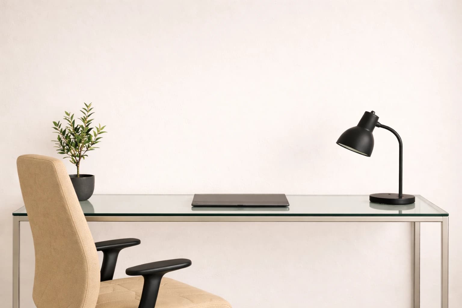 Schreibtisch mit Glasplatte, geschlossenem Laptop mittig, schwarzer Lampe rechts, Pflanze links und beigem Stuhl vor weißer Wand.