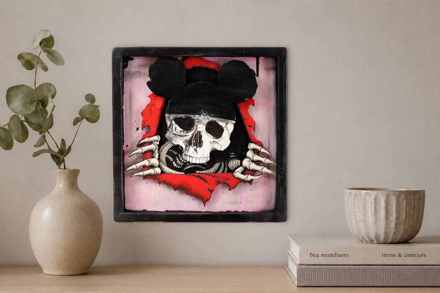 Totenschädel mit schwarzen Mausohren und roten Haaren vor zerrissenem rosa Hintergrund im schwarzen Rahmen, als Kunstwerk an der Wand eines hellen Raums.