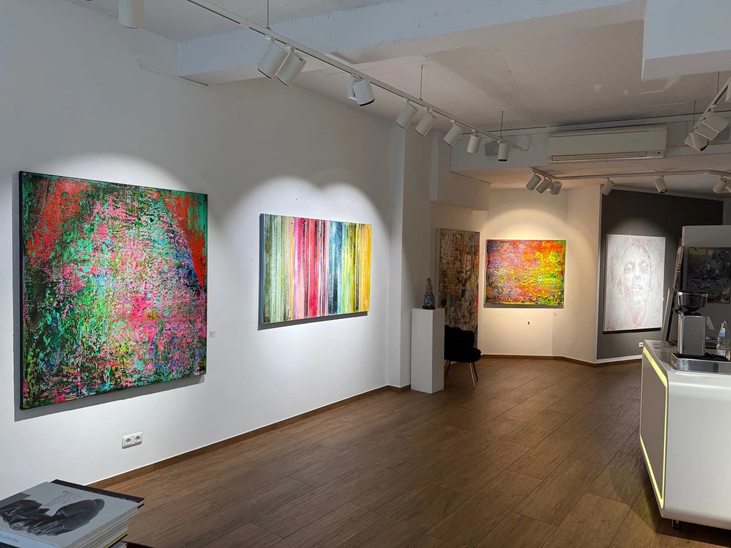 Galerieraum mit vier farbigen Bildern an weißen Wänden und Holzfußboden, Theke rechts.