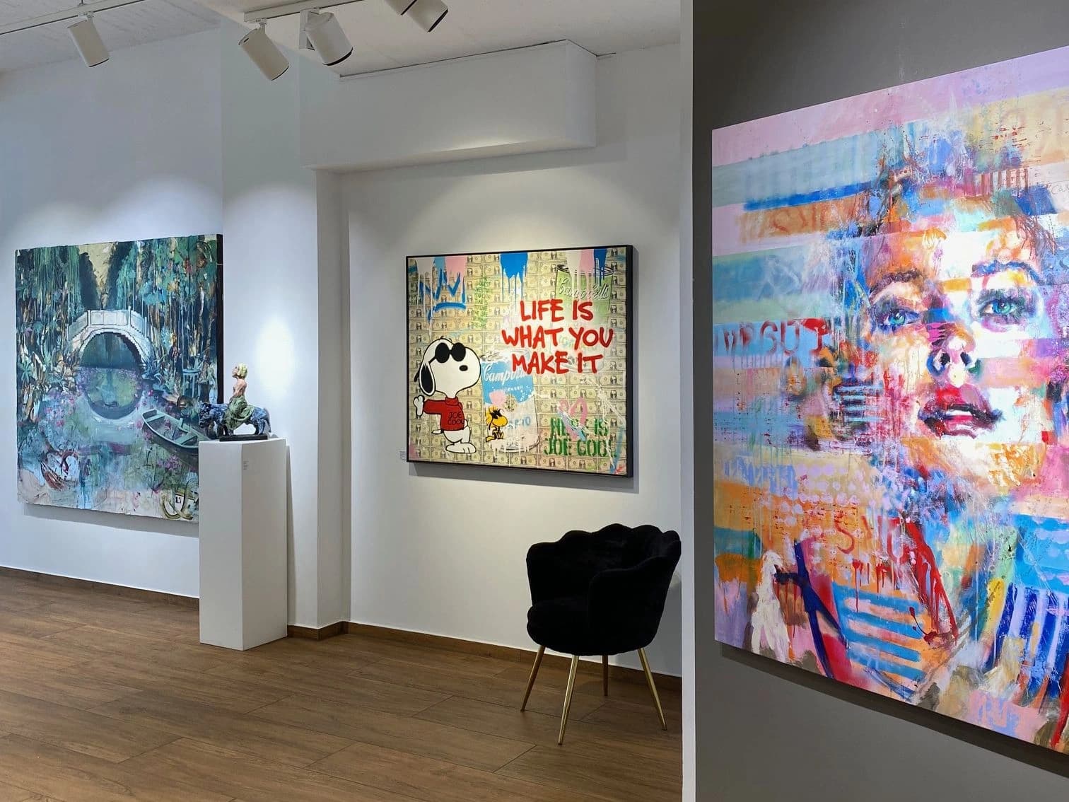 Galerie mit drei farbigen Gemälden an weißen und grauen Wänden, Holzfußboden und schwarzem Sessel.