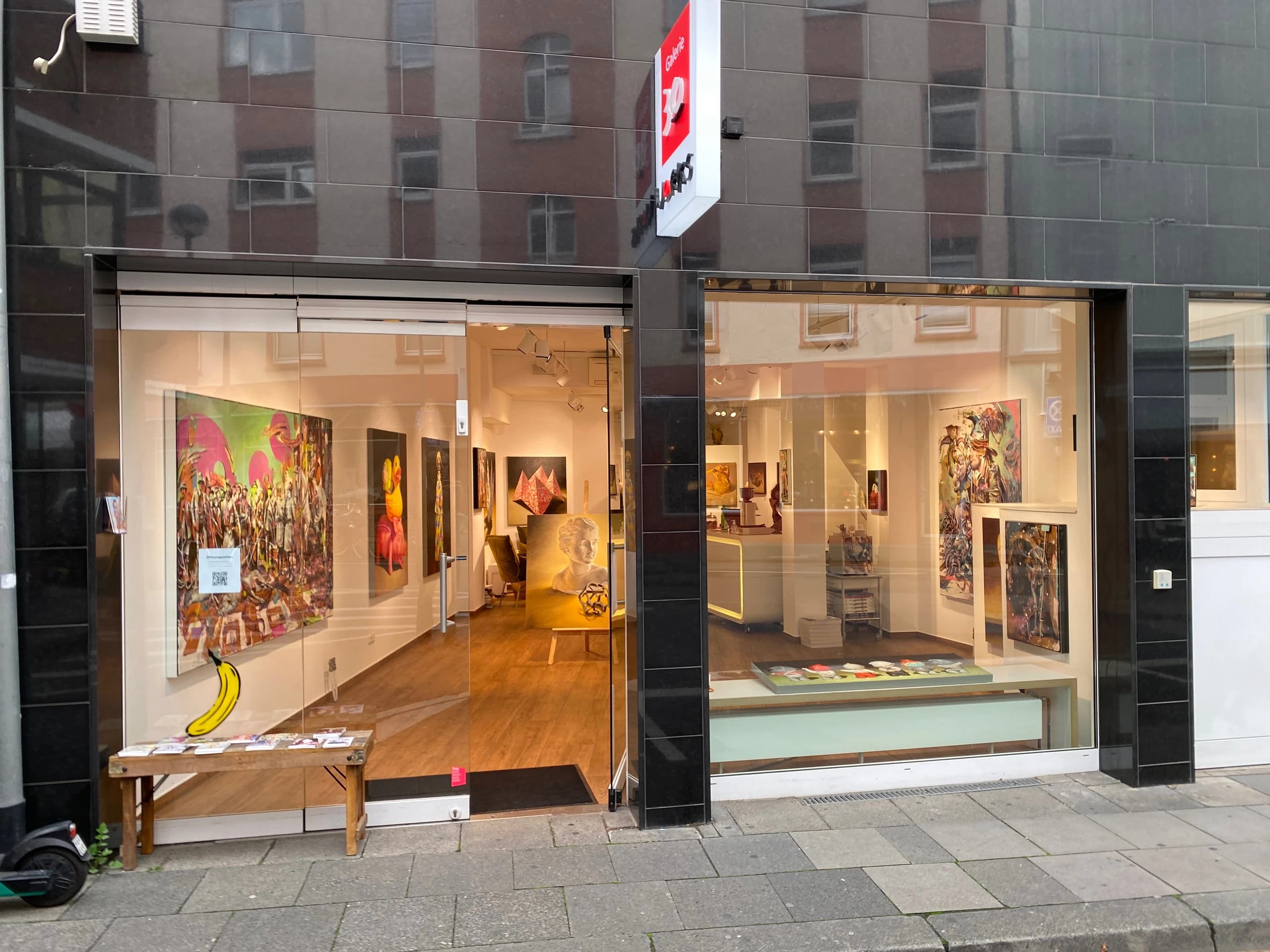 Kunstgalerie mit offenen Schaufenstern, Bildern an den Wänden, Holzboden und Bank vor dem Eingang.