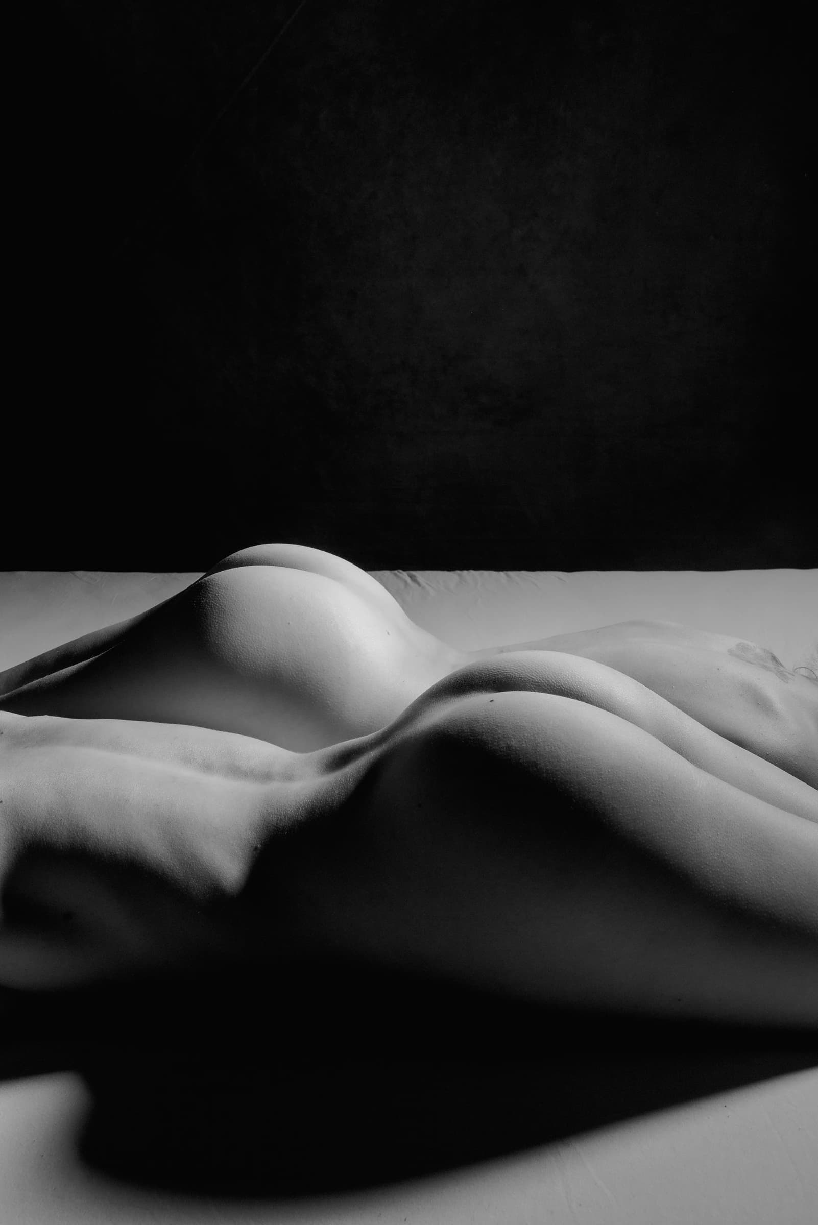 Schwarze Fotografie von zwei nackten Rücken auf einem hellen Tuch vor dunklem Hintergrund.