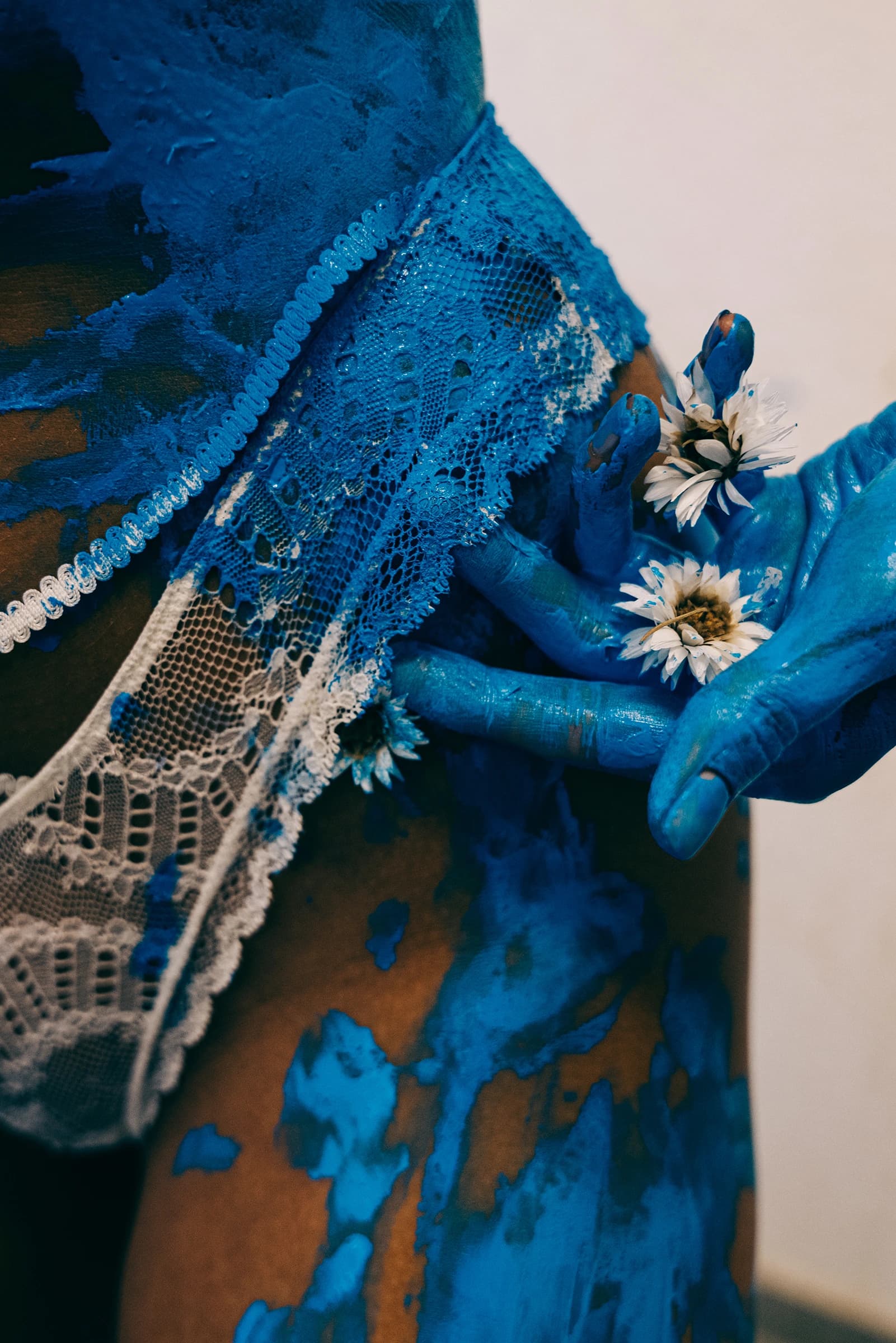 Blaue Hände halten weiße Blumen an einem mit blauer Farbe bespritzten Oberkörper mit Spitze.