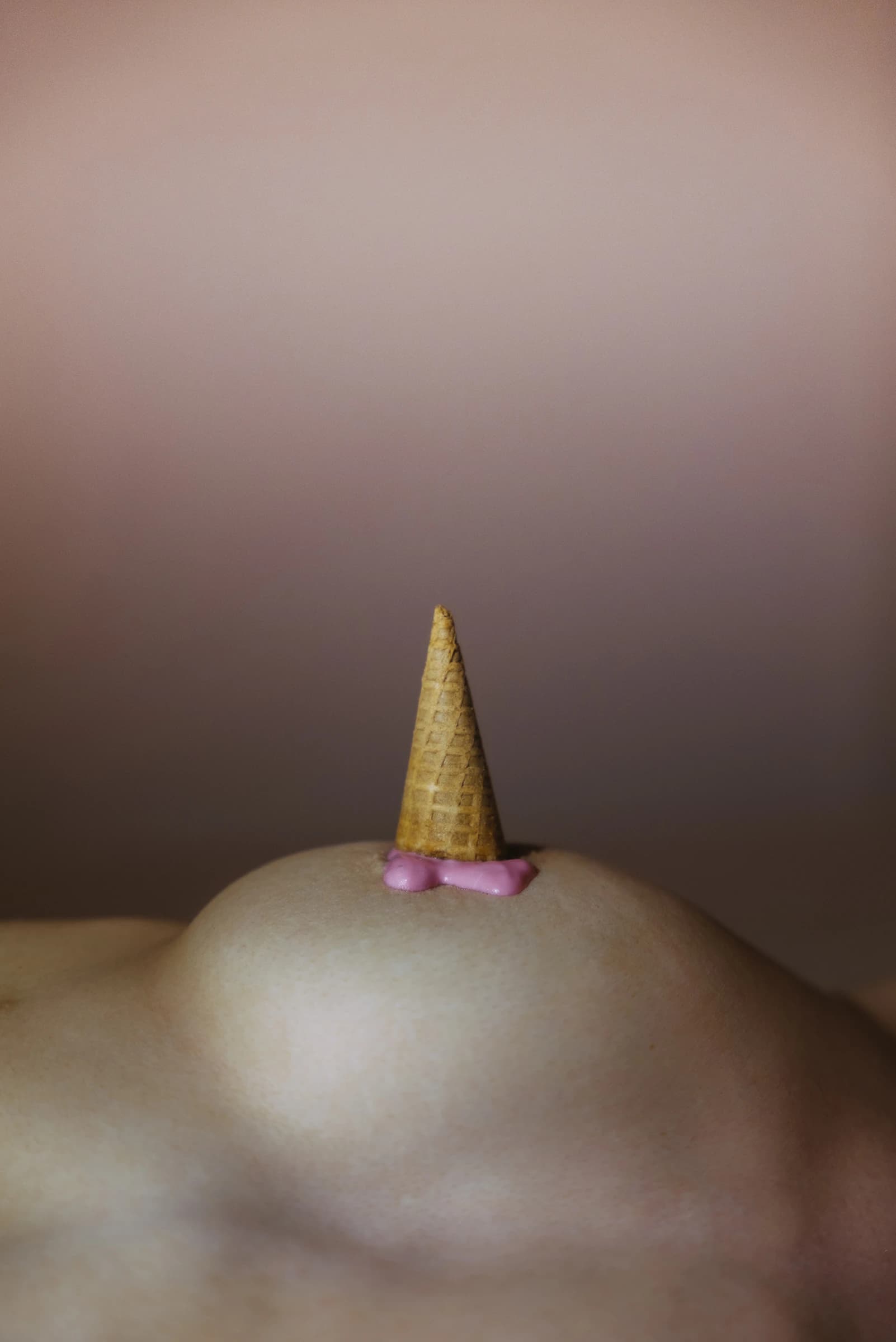 Ein nackter Oberkörper mit aufgesetzter Eistüte und rosa Creme vor braunem Hintergrund.